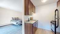 This single-floor unit is thoughtfully laid out to maximize functionality within a compact footprint. The galley-style kitchen is a standout feature, equipped with rich warm-toned wood cabinetry extending to the upper walls, granite-style countertops, a subway tile backsplash, a double-basin sink, a gas range, and a full-size refrigerator in a dark finish. Light wood-tone flooring runs consistently throughout the unit, lending a cohesive and clean aesthetic to the entire space.

The sleeping area is bright and simply designed, with neutral white walls and the same light hardwood-style flooring. A set of full-height built-in closet doors provides generous storage along one wall. A large sliding glass door floods the room with natural light and offers direct access to an outdoor area, delivering views of the surrounding neighborhood.

The bathroom is neatly appointed with a wood-framed vanity mirror, a white basin sink set into a clean countertop, matching warm wood cabinetry below, and a tub-shower combination. Wood-look tile flooring ties the bathroom's design to the rest of the unit. Throughout the space, the design choices are consistent and practical, creating a functional and cohesive living environment.