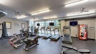 This fitness center is a comprehensive, single-floor facility designed to support a wide range of workout routines. The cardio zone is a highlight of the space, featuring a row of treadmills and elliptical trainers positioned along a large window wall that brings in ample natural light and offers views of surrounding greenery — an inviting backdrop for endurance training. Each treadmill is equipped with an individual mounted screen, and additional flat-screen TVs are positioned throughout the room for entertainment during workouts. The strength training area is equally well-appointed, with a multi-station cable and weight machine, a functional trainer, and a dedicated free weights section. The free weights area includes a full rack of dumbbells in varying sizes, kettlebells, and medicine balls, all set in front of a large wall mirror with a ballet-style bar, making the space functional for both traditional lifting and dynamic training. A heavy punching bag on a stand adds a boxing and functional fitness element to the layout. Instructional fitness posters are displayed on the walls throughout, and a water fountain and vending machine provide added convenience. Exposed ductwork runs along the ceiling, lending an industrial character to the otherwise clean, neutral-toned interior. The rubber flooring throughout ensures safety and durability across all workout zones.