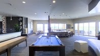This contemporary single-floor amenity space is designed for recreation and social gathering, offering a well-appointed layout filled with natural light streaming through large windows and sliding glass doors that open to an outdoor terrace. At the center of the room stands a full-size pool table with a blue felt surface, complemented by a shuffleboard table nearby, making it a hub for game-day entertainment. A stylish fireplace anchors one wall, adding warmth and character to the lounge atmosphere. The bar area features a curved counter with high-top seating and pendant lighting overhead, creating an inviting spot for casual dining or conversation. A long communal dining table with bar-height chairs provides additional seating toward the back of the space. The room is finished with recessed ceiling lighting, neutral-toned carpeted flooring with subtle linear detailing, and textured walls accented by artwork. Structural columns punctuate the open floor plan, reinforcing the industrial-modern aesthetic. The open sightlines throughout the space and the seamless connection to the outdoor area enhance the overall sense of openness and versatility, making this an ideal multifunctional amenity lounge.