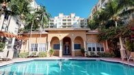 This inviting amenity space showcases classic Mediterranean Revival architecture, immediately recognizable by its warm golden stucco façade, distinctive terracotta tile roof, and graceful arched colonnades that frame the entrance. The single-story clubhouse building serves as the centerpiece of the outdoor amenity area, its elegant arched entryway accented by decorative columns and flanked by large shuttered windows that allow natural light to filter through. French doors at the main entrance invite residents into the interior space. The exterior is beautifully landscaped with tall swaying palm trees and vibrant tropical foliage, including flowering plants that add splashes of color against the warm-toned building. A generously sized swimming pool takes center stage in front of the clubhouse, its crystal-clear turquoise water shimmering beneath an abundant Florida sun. A tiled pool deck surrounds the water and provides space for poolside seating. The setting is completed by a backdrop of a multi-story residential building with private balconies, reinforcing the resort-like character of the community. The overall ambiance is one of relaxed, sun-drenched elegance befitting a tropical retreat.