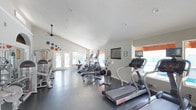This fitness center offers a thoughtfully designed workout space that is both functional and inviting. The room features vaulted ceilings that lend a sense of openness, complemented by recessed lighting and a ceiling fan that help maintain a comfortable environment during exercise. Large windows and glass-paneled doors along the far wall flood the space with natural light while providing views to the outdoors, creating an uplifting atmosphere.

The flooring is a durable dark grey material well-suited for high-traffic athletic use. A row of cardio machines — including treadmills, ellipticals, and a rowing machine — are arranged to maximize the available floor space, allowing for easy navigation between equipment. Wall-mounted televisions are positioned for convenient viewing during workouts.

Adding a touch of personality, colorful circular dot graphics in teal, orange, and red accent the white walls, giving the space a modern and energetic feel. The overall design is clean and minimalist, keeping the focus on functionality. With ample room to move and a bright, airy ambiance, this fitness center provides a welcoming and well-organized environment suited for a range of cardiovascular and general fitness routines.