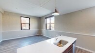 This single-floor urban loft-style unit masterfully combines industrial heritage with contemporary design. The open-concept living and kitchen area serves as the heart of the space, where whitewashed exposed brick walls and a striking pendant light create a warm, characterful ambiance. Large black-framed windows flood the area with natural light and frame views of the surrounding cityscape. The kitchen island features a sleek quartz countertop with a stainless steel sink, complemented by dark cabinetry, wood-look flooring, and a window that further connects the cooking space to the urban surroundings.

The bedrooms offer a quieter retreat, with plush carpeted floors and continued exposed brick and raw concrete accent walls that reinforce the industrial aesthetic. Each room benefits from natural light through well-placed windows. A spacious walk-in closet provides generous storage with built-in shelving units and double hanging rods, offering excellent organizational capacity.

The bathroom is finished with modern fixtures, including a dark wood vanity topped with a white countertop, a framed mirror, a glass-enclosed shower, and wood-look tile flooring. A bold, dark-stained entry door with a brushed steel frame rounds out the unit, making a strong first impression. Throughout, the space balances raw industrial elements with clean, modern touches.