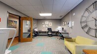 Located within the University Hills Blvd Business Center in Toledo, Ohio, this efficiently designed single-room office space offers a well-organized, multi-functional environment suited for both focused work and collaborative interaction. The space is finished with a drop tile ceiling equipped with recessed fluorescent lighting panels that provide consistent, even illumination throughout.

The room is divided into two functional zones. Along one wall, a long desk surface accommodates multiple workstations with ergonomic task chairs, a desktop computer, printer, and wall-mounted supply station, making it well-equipped for day-to-day office tasks. Framed artwork arranged above the desk adds a polished, professional touch to the gray-painted walls.

The lounge area features two yellow upholstered armchairs arranged around a wooden side table with small decorative plants, anchored by a striking oversized metal clock mounted on the wall. A high-top round table with metal stools and a wall-mounted whiteboard provide additional space for informal meetings or brainstorming sessions. The patterned carpet tiles — featuring a neutral base with teal accent squares — tie the zones together visually, contributing to a modern, cohesive aesthetic throughout the space.