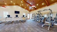 Located at 51 Cohen Walker Dr in Warner Robins, Georgia, this fitness center is a well-appointed, single-floor facility designed to support a comprehensive workout experience. The space is immediately distinguished by its dramatic vaulted ceiling clad in warm natural wood, which rises to impressive heights and lends an open, airy quality to the interior. Recessed lighting is embedded throughout the ceiling, complemented by striking red pendant barn lights that add a bold, stylish accent to the overall aesthetic.

The flooring throughout the main workout area consists of durable, patterned carpet tiles, providing comfort and traction underfoot. A wall-mounted flat-screen television is positioned for easy viewing during cardio sessions. Large mirrors line one wall, enhancing the sense of space and allowing users to monitor their form during strength training.

The cardio zone is equipped with multiple treadmills and additional cardio machines, while the strength training area features cable machines, weight benches, a rack of kettlebells, and a selection of dumbbells. Isolated strength machines are also present for targeted muscle training.

Natural light filters in through windows fitted with classic plantation shutters, and easy access is provided through a warm wood-framed glass door. The overall ambiance is clean, functional, and inviting — a purpose-built space well-suited for daily fitness use.