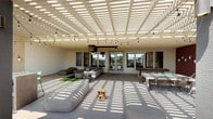 This covered outdoor terrace is designed as a multifunctional social space, anchored by a white slatted pergola that casts striking geometric light patterns across the floor throughout the day. String lights are strung beneath the pergola, adding ambiance for evening gatherings. The terrace is divided into distinct activity zones that flow seamlessly into one another.

A lounge area features generous sectional seating in neutral gray tones, accented with yellow and green throw pillows, arranged around a concrete drum coffee table on a turf rug. A ceiling-mounted projector serves this zone, making it suitable for outdoor screenings. Large glass windows and doors connect this area to an interior space beyond.

The dining zone includes a substantial rustic wood table surrounded by sleek black wire chairs, centered with a fresh greenery centerpiece. Nearby, a classic wooden shuffleboard table provides additional entertainment. A terracotta-red accent wall is adorned with a curated row of hanging wall planters, adding a decorative and organic touch.

The open end of the terrace features glass railings, allowing unobstructed views of the surrounding residential community and landscaping, filling the space with natural light and an airy, connected atmosphere.