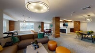 This generously proportioned amenity space is designed to accommodate a variety of social and recreational activities across one thoughtfully arranged floor. The open layout is anchored by striking wood-clad columns that provide visual structure while maintaining a sense of connectivity throughout the space.

The main lounge area features comfortable sectional seating, swivel chairs, and leather ottomans arranged around a central coffee table, with a wall-mounted television and large windows offering ample natural light. Adjacent to this is a game area equipped with an air hockey table and casual seating, bathed in light from surrounding windows.

A dedicated wet bar and kitchenette area stands out with dark wood cabinetry, quartz countertops, a stone tile backsplash, a built-in sink, and a mounted television—finished above with a warm wood-paneled ceiling detail. This area flows seamlessly into a dining and collaborative zone with tables and colorful chairs.

A glass-enclosed private meeting or co-working room with black-framed doors offers a more focused environment. Throughout the space, recessed lighting, pendant fixtures, and patterned carpeting contribute to a modern, welcoming ambiance suited for both relaxation and social gatherings.