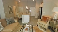 This single-floor space showcases an open-concept layout that seamlessly connects the living area and kitchen, creating a cohesive and functional environment. The kitchen serves as a focal point, featuring a white central island with a granite countertop, a stainless steel refrigerator, and a stainless steel range. White cabinetry provides ample storage, while pendant lighting suspended from the soaring ceilings adds a refined touch over the island. The polished concrete flooring extends throughout, lending an industrial-chic aesthetic that complements the neutral color palette.

The living area is furnished with cream-toned seating, including a sofa and an accent chair with warm-toned throw pillows, arranged around a glass-top coffee table atop a textured area rug. A gallery wall with framed artwork adds visual interest near the kitchen, enhancing the welcoming ambiance. The high ceilings contribute to an airy, expansive feel throughout the space. An entry area is visible near the kitchen, with a door leading to additional rooms. Overall, the space balances modern design elements with warm, comfortable furnishings.