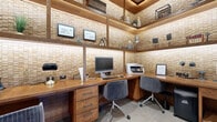 This thoughtfully designed business center offers a compact yet highly functional workspace that blends rustic warmth with professional utility. The room is defined by its striking three-dimensional stone-tile accent walls in warm sand and beige tones, which wrap around the interior and lend rich texture and depth to the space. Dark wood framing contrasts beautifully against the lighter stone, creating a refined, lodge-inspired aesthetic throughout.

Built-in wooden shelving lines the upper perimeter of the room, illuminated by warm LED strip lighting that casts a soft, inviting glow. The shelves display decorative items, books, and greenery, adding personality without sacrificing function. An L-shaped wooden workstation along two walls provides ample desk space, equipped with a desktop computer, printer, and multiple power outlets to support a full range of business needs. Paired with comfortable upholstered task chairs, the setup accommodates multiple users simultaneously.

A cushioned wooden bench seat along one wall offers an additional area to sit and work or wait, accompanied by framed artwork and decorative wall pieces that enhance the curated, welcoming atmosphere. Large-format neutral floor tiles ground the space with a clean, contemporary finish. A bold oversized wall clock near the glass-panel entry door adds a striking focal point. The room is accessed through black-framed glass doors, connecting it seamlessly to the surrounding facility.