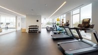 Located at 10 Waverly Street in Boston, MA, this modern fitness center offers a well-appointed, single-floor facility designed to support a comprehensive workout experience. The main exercise area is spacious and open, finished with dark rubber flooring that provides both durability and comfort underfoot. Floor-to-ceiling windows line the perimeter, flooding the space with abundant natural light and offering elevated views of the surrounding Boston skyline and treetops, creating an energizing atmosphere.

The cardio zone is equipped with multiple treadmills featuring integrated screens, as well as elliptical machines, all positioned to take advantage of the expansive outdoor views. A dedicated free weights section includes a full dumbbell rack, kettlebells, plyo boxes, and an adjustable weight bench, catering to strength training needs. A variety of accessories such as exercise balls, resistance bands, and other fitness tools are also available throughout the space.

A separate studio room, distinguished by its teal-blue accent walls and warm wood shelving, houses cycling equipment and a large wall-mounted display screen — ideal for immersive, instructor-led workouts. Recessed and linear LED lighting throughout ensures consistent illumination across all areas. The overall design is clean and contemporary, combining white brick accent walls with a neutral palette to create a professional and motivating environment.