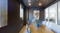 This single-floor space includes a well-appointed meeting or conference room that combines modern design with a functional layout. The room is characterized by deep charcoal-toned walls that create a focused and professional atmosphere. A warm-toned oval wooden table serves as the centerpiece, surrounded by contemporary white chairs that provide a clean contrast to the darker surroundings. Recessed ceiling lighting provides consistent, even illumination throughout the room, complemented by natural light filtering in through a horizontal window on one wall. A framed art piece depicting a classic streetscape adds a distinctive decorative touch to the space. A potted aloe plant atop the table introduces a natural element, softening the room's overall aesthetic. Light wood-toned flooring runs throughout, adding warmth to the otherwise cool color palette. A glass partition or doorway visible on one side of the room suggests connectivity to adjacent areas, enhancing the sense of openness. Overall, the space balances a sleek, modern design sensibility with practical meeting functionality.