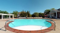 Located at 5353 Wilcox Rd in Dublin, Ohio, this outdoor amenity space centers around a generously sized swimming pool that serves as a standout feature of the property. The pool is finished in a clean, bright aqua blue and is bordered by a classic red brick surround that adds a warm, traditional aesthetic to the space. A wide pool deck extends along the perimeter, accommodating a full row of lounge chairs and chaise seating ideal for sun-bathing and relaxation. The area is framed by tall, mature evergreen and deciduous trees that provide natural privacy and shade along the edges of the space, creating a pleasant, resort-like atmosphere. The surrounding landscape includes well-maintained greenery, contributing to the serene outdoor environment. A red patio umbrella is visible in the background, suggesting an additional shaded seating area for casual gatherings. The property's multi-story residential building is visible beyond the treeline, giving context to the communal nature of this outdoor amenity. Under open skies, this pool area offers a bright, inviting retreat well-suited for warm-weather enjoyment by residents of the community.
