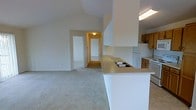 This single-floor residence offers an open and airy layout designed for comfortable everyday living. The kitchen is positioned at the heart of the home, featuring a peninsula counter with a double-basin sink, warm wood-toned cabinetry, a white refrigerator, and a dishwasher. The peninsula creates a natural divide between the kitchen and the adjoining living area while maintaining an open, connected feel throughout.

The living area is finished with light-toned wall-to-wall carpeting, contributing to a warm and quiet atmosphere. Vaulted ceilings rise overhead, adding a sense of openness and vertical space that enhances the overall sense of scale. Neutral white walls throughout keep the interior bright and versatile.

A hallway visible from the main living space leads to additional rooms beyond, including what appears to be a bedroom with a closet, suggesting a practical and well-organized floor plan. Flush-mount and fluorescent lighting fixtures provide consistent illumination across the space.

Overall, the unit presents a clean, functional design with an emphasis on open sightlines, practical storage, and a cohesive neutral palette that makes it easy to personalize and furnish.