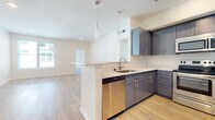 This well-appointed single-floor unit offers a clean, contemporary design throughout. The open-concept layout connects the kitchen and living area seamlessly, creating a spacious and functional flow. The kitchen is a standout feature, equipped with slate-gray shaker-style cabinetry, brushed nickel hardware, white quartz countertops, a subway tile backsplash, and a full suite of stainless steel appliances including a refrigerator, range, microwave, and dishwasher. A peninsula with pendant lighting serves as both a prep space and a casual dining area, anchoring the kitchen's connection to the main living space. Light hardwood flooring runs consistently throughout, lending warmth and continuity to every room. The living area is generously proportioned, with recessed lighting and a large window that draws in natural light. The entryway is bright and inviting, featuring a glass-panel front door with a transom window above, flooding the foyer with additional natural light. The bedroom is a comfortable, well-lit room with a ceiling fan and a window overlooking the surrounding neighborhood. The bathroom is finished with matching gray cabinetry, a double-sink vanity with white countertops, a large mirror, and brushed nickel fixtures, maintaining the cohesive modern aesthetic found throughout the unit.