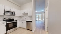 This 450-square-foot one-bedroom apartment at Legacy Village Apartment Homes (Unit E1B) offers a well-designed single-floor layout that maximizes functionality. Light-toned hardwood-style flooring runs throughout, creating a cohesive and airy feel across all rooms. The combined kitchen and living area forms the heart of the unit. The kitchen is outfitted with stainless steel appliances — including a refrigerator, range, microwave, and dishwasher — complemented by white shaker-style cabinetry, light countertops, and a distinctive hexagonal tile backsplash. The living area features a ceiling fan and large French doors that open to a private balcony, filling the space with natural light and outdoor views. The bedroom is a calm, neutral-toned retreat with wall sconce lighting and ample wall space. The bathroom includes a full tub-shower combo with white tile surround, a vanity, and matching light flooring. A dedicated laundry area houses a full-size GE washer and dryer with overhead shelving. The unit is finished with neutral beige walls, white trim, and a keypad-enabled entry door, combining modern convenience with clean, contemporary design.