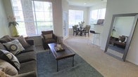 This single-floor space showcases an open-concept layout that connects the living and dining areas in a cohesive, functional design. The living area is anchored by plush carpeting and features a large area rug in a muted blue-grey tone, complementing the neutral wall colors. A dark wood coffee table serves as the centerpiece, accompanied by comfortable seating including an upholstered armchair with decorative pillows.

Large windows throughout flood the space with generous natural light, offering views of surrounding greenery and creating an airy atmosphere. A glass-paneled French door provides additional light and access to the exterior.

The dining area transitions naturally from the living space and is set beneath a bay-style window arrangement, enhancing its bright and welcoming character. A round dark wood dining table is surrounded by cushioned chairs, creating an intimate setting for meals.

A breakfast bar with modern bar seating connects the dining area to an adjacent kitchen space, adding a casual dining option and reinforcing the open flow of the floor plan. Crown molding details along the ceilings add a refined architectural touch throughout.