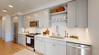 This efficiently designed one-bedroom, one-bathroom residence makes excellent use of its single-floor layout, combining modern finishes with a clean, cohesive aesthetic throughout. The galley-style kitchen is a standout feature, showcasing light gray shaker cabinets with gold hardware, white subway tile backsplash, quartz countertops, stainless steel appliances including a gas range and built-in microwave, and open shelving for added storage and display. Light hardwood flooring runs throughout, connecting each room seamlessly. The living room is bathed in natural light through large windows with sheer curtains, offering urban views and a welcoming, relaxed atmosphere. A ceiling fan adds functional comfort to the space. The bedroom is a calm retreat, featuring elevated clerestory windows that allow soft, diffused light while maintaining privacy. A dedicated closet provides convenient storage. The bathroom is finished with gray shaker vanity cabinetry, a backlit mirror, white tile walk-in shower with a glass enclosure, and neutral tile flooring, creating a spa-like feel. A light-filled hallway connects the rooms, with louvered closet doors adding a classic architectural detail. The overall design is modern, cohesive, and thoughtfully appointed.