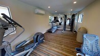 This dedicated fitness center is thoughtfully designed to support a comprehensive workout experience. The space is laid out along a single open floor, maximizing the available area for a variety of exercise equipment. The flooring features a multi-toned striped carpet that adds a sense of energy and directionality to the room.

Cardio equipment is prominently featured, including multiple treadmills and an elliptical trainer, all positioned to take advantage of natural light streaming in through the windows along the side wall. A multi-station cable and strength training machine is positioned toward the far end of the room, offering users options for resistance and weight-based workouts.

A full-length mirror spans a portion of one wall, allowing users to monitor their form during exercise while also visually expanding the space. The ceiling is fitted with recessed lighting, providing bright, even illumination throughout. A wall-mounted split air conditioning unit ensures a comfortable climate regardless of the season.

The overall ambiance is clean, functional, and professional, reflecting a facility designed with active use in mind. The neutral wall tones and practical layout contribute to a focused, distraction-free environment well suited for daily fitness routines.
