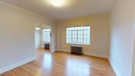 This single-floor space exudes classic architectural charm with a thoughtfully preserved pre-war aesthetic. The main living area is open and airy, showcasing beautifully maintained hardwood floors that catch and reflect natural light throughout the day. Large, multi-pane windows dominate the walls, flooding the interior with an abundance of diffused natural light and lending the space an open, welcoming atmosphere.

Elegant crown molding frames the ceilings, adding a refined architectural detail that speaks to the building's heritage craftsmanship. Cast-iron radiators beneath the windows are a hallmark of classic construction and provide a functional yet characterful design element consistent with the period architecture.

A doorway connects the main living area to an adjacent room, which features its own multi-pane window and a pendant light fixture, offering a versatile space that can serve a variety of purposes. The neutral, warm-toned walls complement the natural wood flooring and contribute to a calm, understated ambiance throughout.

Overall, this space balances timeless architectural details with a clean, move-in-ready condition, making it an inviting environment that honors its historic roots while offering a bright and functional layout.