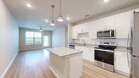 This thoughtfully designed single-floor residence offers three bedrooms and two bathrooms across a comfortable and functional layout. The open-concept kitchen and living area serves as the heart of the home, featuring crisp white cabinetry, granite countertops, a subway tile backsplash, and a full suite of stainless steel appliances including a refrigerator, range, microwave, and dishwasher. A central kitchen island with a matching granite surface provides additional prep space and casual seating. Pendant lighting with brushed gold fixtures illuminates the kitchen, while recessed lighting adds a clean, modern touch throughout. Light wood-look flooring runs through the main living areas, lending warmth and continuity to the space. The living room benefits from direct outdoor access through a glass door flanked by large windows, inviting natural light and exterior views. A ceiling fan enhances comfort in this area. The bedrooms are carpeted for added coziness and are each equipped with windows that frame outdoor views. The generously sized primary bedroom includes a closet for ample storage. Both bathrooms are finished with granite-topped vanities and wood-look flooring; one features a tub-shower combination, while the other offers a streamlined vanity setup. The overall aesthetic is clean, neutral, and move-in ready.
