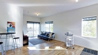 This single-floor space showcases a clean, contemporary design with an emphasis on open living and natural light. The main living area features warm hardwood flooring that extends throughout, reflecting sunlight and enhancing the sense of spaciousness. Crisp white walls provide a neutral backdrop that complements the modern aesthetic.

Large windows and glass sliding doors flood the interior with abundant natural light while offering views of the surrounding outdoor area and direct access to a private balcony. The balcony extends the living space outdoors and provides an open-air retreat.

The living area is thoughtfully laid out, accommodating a full seating arrangement including a navy sofa with accent pillows and a wire-frame accent chair, anchored by a low-profile area rug. A rolling utility shelf adds a functional, industrial touch to the space, while a dark wood media console grounds one end of the room.

The overall ambiance is bright, minimal, and modern — ideal for those who appreciate clean lines, natural materials, and a well-lit interior. The combination of hardwood floors, generous window placement, and access to outdoor space makes this a practical yet stylish single-level residence.