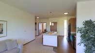 This single-floor space is designed with an open-concept layout that integrates the kitchen and living areas into one cohesive environment. The kitchen serves as the focal point, anchored by a white central island with a light-toned countertop that doubles as a breakfast bar with seating. The island is equipped with a sink and is illuminated by pendant lighting suspended from above, adding a modern touch to the space.

The kitchen is outfitted with dark stainless steel appliances, including a refrigerator and range, and is complemented by warm wood-toned upper cabinetry along the perimeter wall. Hard flooring runs through the kitchen and dining zone, providing a practical and clean aesthetic, while plush carpet covers the adjoining living area, creating a clear visual distinction between the two spaces.

The ceilings are standard height and finished in white, contributing to a bright and clean atmosphere. Neutral-toned walls throughout allow for versatile decorating options. Doorways visible in the background suggest additional rooms beyond, enhancing the functional layout of this well-organized, single-level space.