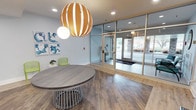 This single-floor space is thoughtfully designed with a clean, modern aesthetic that balances functionality and style. The welcoming entry lobby sets the tone immediately, featuring warm wood-look flooring, a striking oversized globe pendant light in gold and white tones, and a sculptural round table with slatted base. Blue abstract wall art and lime green accent chairs add a vibrant, contemporary touch, while floor-to-ceiling glass doors provide an open connection to the exterior.

Adjacent to the lobby, a glass-partitioned room with light wood framing houses teal accent chairs and a decorative circular wall mirror, offering a semi-private seating or waiting area with clear sightlines throughout the space. The flooring transitions subtly between sections, adding visual interest.

A bright communal area features a long white bar-height island with warm cognac-toned counter stools beneath a large rectangular pendant light. Floor-to-ceiling windows flood this zone with natural light and offer views of an outdoor terrace.

A wide, well-lit corridor extends through the space, finished with the same light hardwood flooring and crisp white walls. A built-in illuminated glass display niche adds a refined decorative element. Recessed and flush-mount ceiling lights ensure consistent, clean illumination throughout.