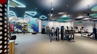 This expansive, single-floor fitness center offers a thoughtfully designed layout divided into distinct training zones, each with its own character and purpose. The main gym area is immediately striking, with a dark, dramatic ceiling accented by geometric LED light installations that cast a vibrant, energizing glow throughout the space. Suspended teal acoustic panel fixtures add both visual interest and sound management overhead, while resilient rubber flooring provides comfort and durability underfoot.

The cardio zone features a full row of treadmills and upright bikes, complemented by stair climbers and rowing machines. The strength training area is equipped with a comprehensive selection of cable machines, plate-loaded equipment, free weights, barbells, and power racks, all set against bold, large-scale pop-art murals that animate the walls with dynamic athletic imagery.

A separate functional training room offers a calmer, more intimate setting finished with light hardwood flooring, full-length mirrors, pendant lighting, and a well-stocked multi-purpose equipment rack loaded with medicine balls, resistance bands, and foam rollers — ideal for stretching, mobility work, or personal training sessions.

Throughout the facility, the design balances high energy with purposeful organization, creating a professional and visually engaging environment suited to a wide range of fitness pursuits.