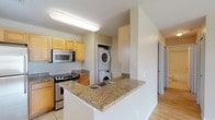 This single-floor residence offers a well-organized layout with distinct living, sleeping, and utility zones. The open-concept kitchen serves as a central hub, outfitted with light maple cabinetry, granite-style countertops, and stainless steel appliances including an electric range with overhead microwave. A peninsula with an integrated sink and additional counter space extends into the living area, creating a natural divide between the kitchen and main living space. A stacked washer and dryer unit is conveniently housed in a dedicated alcove directly off the kitchen.

The living area features light wood-style flooring and a large window that allows natural light to fill the space and offers an outdoor view. A front entry door opens directly into this area, providing a straightforward and functional flow throughout the unit.

The bedrooms are carpeted and finished in neutral tones, each offering closet access. One bedroom includes a window with outdoor views and a bifold closet door. Both bathrooms are tiled and share a consistent design featuring maple vanity cabinetry, granite-style countertops, a large mirror, and a tub-shower combination, providing a cohesive, practical finish throughout the space.