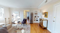 This refined single-floor residence exudes a timeless charm, thoughtfully designed with attention to detail and classic architectural finishes. Warm hardwood flooring flows throughout the space, complemented by crisp white walls and elegant crown molding that lend a polished, cohesive feel to every room.

The living area serves as a welcoming centerpiece, bathed in generous natural light streaming through large windows. A plush area rug anchors the seating arrangement, where a tufted velvet accent chair adds a touch of sophistication. Built-in white shelving lines one wall, offering both functional storage and a curated display space for decorative objects and books. An ornate mirrored dresser and a gold-framed painting enhance the room's refined aesthetic.

A decorative arrangement of dried branches adds an organic, artistic element, bridging the natural and the curated. The open sightline into an adjoining bedroom reveals continued brightness and a consistent design language throughout the home.

Crown molding details, recessed air vents, and carefully considered furnishings reflect a space that balances comfort with elegance. Overall, this residence offers a serene and well-lit environment with classic design elements that create a warm, inviting ambiance ideal for everyday living.
