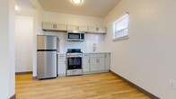 This well-designed one-bedroom, one-bathroom unit offers a practical and modern living space across a single floor. Light wood-style luxury vinyl flooring runs throughout the entire unit, creating a clean, cohesive aesthetic.

The kitchen is a standout feature, equipped with stainless steel appliances including a full-size refrigerator, gas range with oven, and an over-the-range microwave. Gray shaker-style cabinetry with matte black hardware lines the walls both above and below the counter, complemented by a white subway tile backsplash and a single-basin sink with a modern faucet. A window provides natural light to the cooking area.

The bedroom is a comfortable, well-proportioned room featuring multiple windows that allow generous natural light and offer views of surrounding greenery. The bright white walls and light flooring contribute to an airy, open feel.

The bathroom is clean and functional, featuring a full tub-shower combination, a toilet, and a vanity with cabinet storage. Light flooring and white walls maintain the unit's consistent, fresh aesthetic, with a window providing additional ventilation and light.

A hallway connects the rooms efficiently, completing this thoughtfully laid-out unit that maximizes every square foot of available space.