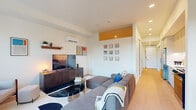 This thoughtfully designed one-bedroom, one-bathroom residence makes excellent use of its single-floor layout, combining modern aesthetics with practical functionality. The living area serves as the heart of the home, featuring light hardwood flooring, recessed lighting, and a wall-mounted mini-split system for year-round climate control. A bold amber-yellow accent panel above the hallway entry adds a striking pop of color, complemented by a curated gallery wall of colorful framed artwork. The living room comfortably accommodates a large sectional sofa, a dark wood media console, and a glass coffee table, creating a stylish and inviting atmosphere. A sliding barn door connects the living space to the private hallway, lending an open, modern touch. The bedroom is a serene retreat, distinguished by a dramatic clerestory window that draws in natural light and treetop views. Warm-toned textiles and wall hangings add character and comfort to the space. The bathroom features a clean, all-white design with a wide vanity, undermount sink, ample drawer storage, and a full-size mirror, reflecting a minimalist yet functional approach. Light hardwood floors carry throughout, unifying the space with a cohesive, contemporary feel.