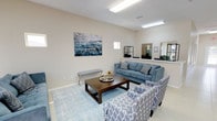 This single-story commercial space functions as a leasing office, thoughtfully designed to provide a comfortable and professional environment for prospective tenants and staff alike. The interior is characterized by neutral beige walls, light-toned large-format tile flooring, and elevated ceilings that lend an open, airy feel throughout.

The main reception and lounge area serves as the focal point of the space, furnished with a plush blue sofa, coordinating patterned accent chairs, and a dark wood coffee table anchored by a light blue area rug. Abstract wall art in muted blue and gray tones adds a decorative touch, complementing the overall color palette. Multiple windows along the front façade allow generous natural light to fill the room, while recessed and flush-mount fluorescent lighting ensures consistent illumination.

An adjacent area features a tall dining-height table with upholstered chairs, ideal for consultations or informal meetings. A long corridor extends toward the rear of the building, lined with additional doors suggesting private offices or meeting rooms. A mirrored wall panel near the main lounge enhances the sense of depth and openness. The layout flows efficiently, balancing functional work areas with welcoming communal spaces suited for a professional leasing environment.