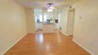 This single-floor space offers a clean, open layout that seamlessly connects the living area to the kitchen, creating a sense of flow and functionality. Warm hardwood-style flooring runs throughout the main living area, adding a welcoming and cohesive aesthetic to the space.

The kitchen is well-appointed with white cabinetry that extends to the upper walls, providing ample storage. A peninsula counter divides the kitchen from the living area, offering additional prep space while maintaining the open feel of the floor plan. White appliances, including a range and refrigerator, complement the light, airy palette of the kitchen. A window above the sink allows natural light to filter in, brightening the workspace.

A ceiling fan with an integrated light fixture is centrally positioned in the main living area, ensuring both comfort and illumination. The neutral-toned walls throughout the space provide a versatile backdrop well-suited to a variety of interior styles. Clean white baseboards frame the flooring and add a crisp, finished look to each room. A hallway leading off the main area suggests additional rooms, contributing to the overall practicality of the layout. The space presents a bright, move-in-ready environment with a straightforward and efficient design.