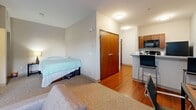 Located at 4830 Cass Ave in Detroit's Midtown neighborhood, this efficiently designed studio unit offers a well-organized single-floor layout that maximizes every square foot. The open-concept kitchen flows directly into the living area, anchored by rich warm-toned hardwood flooring. The kitchen features rich cherry wood cabinetry, granite countertops, a double-basin sink, a built-in dishwasher, and a full-size refrigerator, along with a breakfast bar that doubles as a dining and workspace. The living area is carpeted for comfort and benefits from a large window with horizontal blinds, offering views of the surrounding Midtown Detroit streetscape and filling the room with natural light. A wall-mounted flat-screen television setup and a wall-mounted climate control unit add to the functional amenities of the living space. The sleeping area is integrated within the open floor plan and is delineated by a carpeted zone. The bathroom is appointed with tile flooring, a granite-topped vanity with matching cherry cabinetry, and full fixtures. A set of warm-toned wood double doors provides ample closet storage. The unit also includes the convenience of in-unit laundry. Neutral gray walls throughout lend the space a clean, modern aesthetic.