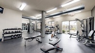 This fitness center offers a spacious and well-organized workout environment designed to support a full range of exercise routines. The main workout area features durable dark rubber flooring throughout, providing both safety and comfort underfoot. Exposed ductwork runs along the ceiling, lending the space an industrial character, while flush-mounted fluorescent lighting ensures the entire area is brightly and evenly illuminated.

The cardio zone is equipped with multiple treadmills and elliptical trainers positioned near large windows, allowing natural light to fill the space and offering views to the outdoors. A stationary spin bike adds further variety to cardio options. The strength training section includes a multi-station cable machine, multiple adjustable weight benches, and free weight equipment, supported by a full-length wall mirror that enhances the sense of openness and assists with form during workouts.

A dedicated stretching and flexibility area features additional benches and open floor space. Wall-mounted shelving provides convenient storage for accessories, and sanitation supplies are readily accessible throughout. A clean corridor connects ancillary rooms, including what appear to be restroom or utility facilities. The combination of ample natural light through large windows and glass doors, modern equipment, and a thoughtfully arranged layout makes this fitness center a functional and inviting space for daily exercise.