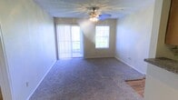 This single-floor residence offers a comfortable and functional layout designed for everyday living. The main living area is a spacious, open room finished with wall-to-wall carpeting in a neutral tone, providing a soft and inviting atmosphere underfoot. A ceiling fan with an integrated light fixture is centrally mounted on the textured ceiling, offering both air circulation and ambient lighting throughout the space.

Natural light flows generously into the room through two key openings: a large sliding glass door dressed with vertical blinds, and a traditional window positioned on the adjacent wall. Together, these features create a bright, airy environment that feels open and welcoming. The sliding door suggests access to an outdoor area such as a patio or balcony, further enhancing the sense of connection to the exterior.

The walls are finished in a clean, light neutral tone, complemented by white baseboards that frame the room with a crisp, tidy appearance. A partial view of an adjacent area with hard flooring is visible, hinting at a seamlessly connected secondary space. Overall, the residence presents a clean, move-in ready environment with practical finishes and an abundance of natural light.