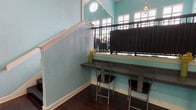 This snack area is a compact yet well-appointed space designed for casual dining and refreshment. The interior is defined by its distinctive light teal walls and rich, dark hardwood flooring, creating a fresh and inviting ambiance throughout. Soaring ceilings with clerestory windows above allow natural light to filter in generously, lending the space an open and airy feel despite its modest footprint.

A built-in counter bar runs along one wall beneath an iron railing, accompanied by metal-framed bar stools that provide a convenient spot for quick bites or light work. A Coca-Cola beverage vending machine is positioned nearby, offering easy refreshment access for visitors. Round tables with metal chairs provide additional casual seating options.

A staircase with a white painted banister is integrated into the layout, adding architectural character to the space. Two gray six-panel doors lead to adjoining areas, including a hallway corridor, ensuring smooth connectivity within the larger facility. Safety features such as a fire extinguisher station and exit signage are clearly visible, reflecting the space's practical, functional design. Decorative touches, including a small floral arrangement and a piece of abstract wall art, add warmth and personality to this efficiently designed snack retreat.