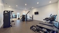 Located at Waterstone Park in Hillsborough, North Carolina, this single-floor fitness center is thoughtfully designed to support a comprehensive workout experience. The facility is organized across several distinct zones, each finished with durable rubber flooring and crisp white walls illuminated by recessed lighting throughout.

The main exercise area is generously proportioned and houses a well-rounded selection of cardio machines, including multiple treadmills, an elliptical, and stationary bikes with interactive digital displays. A cable pulley machine anchors the strength-training zone, while a functional trainer unit offers versatile resistance options for full-body workouts. A wall-mounted flat-screen television provides entertainment during exercise sessions.

A dedicated free weight alcove offers a quiet, focused environment with a tiered dumbbell rack and an adjustable flat bench, ideal for isolated strength training. Mirrors along one wall enhance the sense of space and allow for form checks during exercise.

The facility is accessed via a clean, light-filled corridor with light wood-tone flooring, tall ceilings, and glass-paneled doors that lend an airy, welcoming entry. A water station is conveniently positioned within the main workout area. Natural light filters in through multiple windows fitted with blinds, contributing to a bright and motivating atmosphere throughout the space.