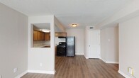 This single-story residence offers a well-organized layout with consistent wood-look flooring running throughout the main living areas, bedrooms, and kitchen, contributing to a cohesive and easy-to-maintain interior. The kitchen is efficiently designed in an L-shaped configuration, featuring warm honey-toned cabinetry with ample upper and lower storage, light granite-style countertops, a stainless steel double sink, and black appliances including a gas range and full-size refrigerator. A pass-through window connects the kitchen to the adjacent living area, enhancing the open feel between spaces. The living and dining areas are open and unobstructed, with neutral wall tones and textured ceilings. Two well-proportioned bedrooms each include ceiling fans with integrated lighting and windows fitted with horizontal blinds that allow natural light while maintaining privacy. One bedroom also features a closet door. The bathroom is neatly appointed with a full tub-shower combination, a wood-vanity with a granite-style countertop, and a large mirror above. White trim and baseboards frame each room cleanly, reinforcing the unit's tidy, move-in-ready presentation throughout.