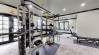 This fitness center offers a spacious, single-floor layout thoughtfully designed to accommodate a comprehensive range of workout needs. The space features striking vaulted ceilings accented by bold black crown molding and exposed beams, lending the room a refined yet functional aesthetic. Recessed lighting and ceiling fans are positioned throughout, ensuring the space remains bright and well-ventilated during use.

Large, black-framed windows and glass-paneled French doors line multiple walls, flooding the interior with abundant natural light and providing views of the surrounding exterior. Full-length wall mirrors further enhance the sense of openness and allow users to monitor their form during exercise.

The strength training area is well-appointed with a multi-functional cable and pulley rig, adjustable benches, free weights, medicine balls, resistance bands, and a dedicated stretching and floor exercise zone with rubber mat flooring. A separate area houses a variety of cardio machines, including treadmills, an elliptical, a stair climber, and a stationary spin bike, all positioned to take advantage of the natural light from the surrounding windows.

The flooring throughout consists of a durable, textured carpet tile that balances comfort and practicality. A wooden storage shelving unit and organized equipment racks contribute to the space's clean, orderly layout. Overall, the fitness center combines a polished architectural design with a comprehensive suite of equipment to support a full-body training experience.