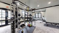 This fitness center offers a spacious, single-floor layout thoughtfully designed to accommodate a comprehensive range of workout needs. The space features striking vaulted ceilings accented by bold black crown molding and exposed beams, lending the room a refined yet functional aesthetic. Recessed lighting and ceiling fans are positioned throughout, ensuring the space remains bright and well-ventilated during use.

Large, black-framed windows and glass-paneled French doors line multiple walls, flooding the interior with abundant natural light and providing views of the surrounding exterior. Full-length wall mirrors further enhance the sense of openness and allow users to monitor their form during exercise.

The strength training area is well-appointed with a multi-functional cable and pulley rig, adjustable benches, free weights, medicine balls, resistance bands, and a dedicated stretching and floor exercise zone with rubber mat flooring. A separate area houses a variety of cardio machines, including treadmills, an elliptical, a stair climber, and a stationary spin bike, all positioned to take advantage of the natural light from the surrounding windows.

The flooring throughout consists of a durable, textured carpet tile that balances comfort and practicality. A wooden storage shelving unit and organized equipment racks contribute to the space's clean, orderly layout. Overall, the fitness center combines a polished architectural design with a comprehensive suite of equipment to support a full-body training experience.