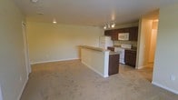 This single-floor space features an open-concept layout that integrates the kitchen and living area into one cohesive environment. The kitchen is equipped with dark brown wood cabinetry, offering ample storage both above and below the countertops. White appliances, including a refrigerator, electric range, and over-the-range microwave, provide practical functionality. A peninsula-style breakfast bar extends into the space, creating a natural division between the kitchen and the adjoining living area while also serving as an informal dining or workspace surface. The kitchen features tile flooring, while the living area transitions to wall-to-wall carpeting, adding warmth and comfort underfoot. Track lighting above the kitchen provides focused illumination, complemented by ceiling-mounted lighting that brightens the living space. The walls are finished in a neutral light tone, contributing to a clean and airy atmosphere throughout. Baseboards are crisp and white, adding a polished finishing detail. The overall layout is straightforward and functional, making efficient use of the available space.