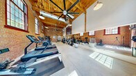 This well-appointed fitness center occupies a generously sized single-floor space that masterfully blends historic industrial architecture with modern amenities. The defining architectural features are the soaring ceilings supported by exposed wooden beam trusses and warm tongue-and-groove wood paneling above, which together create a dramatic and inviting atmosphere. The original exposed brick walls lend an authentic, rustic character throughout the space, while large, multi-pane windows along multiple walls flood the interior with abundant natural light and offer views of the surrounding greenery outdoors.

The polished concrete floors provide a durable, clean surface that complements the industrial aesthetic. Oversized ceiling fans and pendant-style industrial light fixtures hang from the rafters, ensuring excellent airflow and well-distributed lighting. Full-length mirrors along one wall enhance the sense of openness and allow users to monitor their form during workouts.

The fitness floor is comprehensively equipped with a dedicated cardio zone featuring treadmills, elliptical trainers, and stationary bikes, as well as a robust strength-training area outfitted with selectorized machines, a functional trainer cable system, free weights, and benches. Stability balls and additional accessories round out the offerings. A sliding barn-style door and direct exterior access add to the space's unique, converted-building charm.