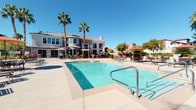 This single-story outdoor amenity space is designed with a resort-inspired aesthetic, offering a comprehensive set of recreational features suited for warm-weather living. The centerpiece is a large, sparkling swimming pool framed by tall palm trees and an expansive sun deck furnished with lounge chairs and market umbrellas, creating an inviting atmosphere for relaxation. A second pool area includes a decorative spa with elegant tile detailing and an adjacent shaded seating nook with planter accents. The clubhouse building, finished in a warm stucco exterior with arched windows and a cupola roofline, anchors the pool courtyard and adds architectural character to the surroundings. A covered outdoor kitchen area features multiple stainless steel gas grills, granite prep countertops, a double-basin utility sink, and a brick-patterned paver floor, providing a fully equipped space for outdoor cooking and entertaining. Adjacent to the kitchen, a covered lounge showcases a floor-to-ceiling natural stone fireplace with a mounted flat-screen television and round patio dining tables with chairs beneath arched openings. A separate patio area features a circular fire pit surrounded by cushioned lounge seating, set among mature trees and lush landscaping, offering a cozy gathering spot for evening use.
