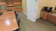 This single-floor professional office space is designed to support a productive and polished work environment. The focal point of the space is a well-appointed meeting or conference room, featuring a substantial wood-toned table surrounded by upholstered striped chairs with wooden frames, offering a comfortable and professional setting for gatherings or discussions. The room is finished with neutral-toned carpeting that lends a quiet, grounded atmosphere throughout.

Built-in wooden cabinetry lines one wall, providing ample closed and open storage, and is accented with tasteful decorative touches. A classic paneled white door serves as the room's entryway, adding a traditional architectural detail that complements the warm wood tones of the furnishings and cabinetry. Adjacent to the main seating area, a dedicated refreshment station features a compact refrigerator and additional cabinetry with a granite-style countertop — a convenient amenity for hosting clients or extended meetings.

The overall ambiance is warm, organized, and functional, with a cohesive color palette of muted greens, warm wood tones, and soft neutrals. The space is well-suited for a professional services environment, offering both utility and a welcoming atmosphere for staff and visitors alike.