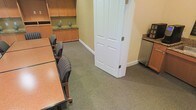 This single-floor professional office space is designed to support a productive and polished work environment. The focal point of the space is a well-appointed meeting or conference room, featuring a substantial wood-toned table surrounded by upholstered striped chairs with wooden frames, offering a comfortable and professional setting for gatherings or discussions. The room is finished with neutral-toned carpeting that lends a quiet, grounded atmosphere throughout.

Built-in wooden cabinetry lines one wall, providing ample closed and open storage, and is accented with tasteful decorative touches. A classic paneled white door serves as the room's entryway, adding a traditional architectural detail that complements the warm wood tones of the furnishings and cabinetry. Adjacent to the main seating area, a dedicated refreshment station features a compact refrigerator and additional cabinetry with a granite-style countertop — a convenient amenity for hosting clients or extended meetings.

The overall ambiance is warm, organized, and functional, with a cohesive color palette of muted greens, warm wood tones, and soft neutrals. The space is well-suited for a professional services environment, offering both utility and a welcoming atmosphere for staff and visitors alike.