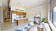 This thoughtfully designed single-floor space showcases a seamless open-concept layout that integrates the kitchen and living areas into one cohesive environment. The kitchen is a standout feature, boasting rich walnut-toned cabinetry, a clean white quartz countertop, and a peninsula with bar seating — ideal for casual dining or entertaining. Stainless steel appliances, including a refrigerator, microwave, and range, provide a polished, professional finish. The cabinetry extends to the ceiling, maximizing storage while drawing the eye upward to the generous ceiling height. Overhead, a combination of track lighting and decorative pendant lights add layers of illumination and visual interest, reinforcing the industrial-modern character of the space. Polished concrete flooring runs throughout, offering both durability and a sleek, contemporary aesthetic that ties the space together. The open living area is bathed in ambient light and offers ample room for flexible furniture arrangements. The overall ambiance is modern and urban, with a clean palette of whites and warm wood tones complemented by metallic accents — creating a space that feels both functional and stylish.