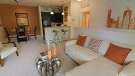 This single-story residence showcases an open-concept floor plan that integrates the living and kitchen areas into one cohesive, functional space. The living area is carpeted and comfortably appointed, anchored by a cream-toned sofa accented with warm orange and patterned throw pillows. A sleek glass-top coffee table with a sculptural metallic base serves as a stylish focal point, dressed with decorative candle holders that add a warm ambiance to the room.

The kitchen is defined by dark cabinetry that contrasts with light granite countertops, giving it a bold, contemporary feel. Black appliances, including a refrigerator and microwave, complement the cabinetry. A breakfast bar with bar-height seating provides a casual dining option and a natural transition between the kitchen and living space. Pendant lighting hangs above the bar area, contributing both task lighting and visual interest.

A hallway visible from the living area leads to additional rooms, suggesting a practical layout throughout. Decorative wall art and warm accent colors tie the space together, creating a cohesive and welcoming interior aesthetic.