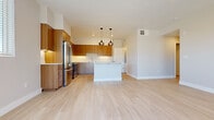 This single-floor residence showcases a contemporary design with a cohesive, neutral palette throughout. The open-concept kitchen serves as the heart of the space, outfitted with warm wood-toned cabinetry extending to the ceiling, a full suite of stainless steel appliances including a French door refrigerator, range, microwave, and dishwasher, and a white countertop island illuminated by a pair of stylish bronze pendant lights. Light wood-look flooring runs through the main living and kitchen areas, lending warmth and continuity.

A central hallway connects the living spaces to the private rooms, where plush carpet flooring provides comfort underfoot. Bedrooms are bright and functional, featuring large windows with horizontal blinds and generous closet space. One room opens directly onto a private balcony through a sliding glass door, offering outdoor access and natural light.

The bathroom is clean and efficiently designed, featuring light wood-toned cabinetry, a white countertop vanity with a large mirror, and a glass-enclosed walk-in shower. Throughout the unit, recessed lighting, white walls, and wide baseboards contribute to a polished, move-in-ready aesthetic.