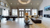 Located at 1 Glastonbury, this refined single-floor clubhouse space is designed to impress with its upscale finishes and thoughtful layout. The main lounge area commands attention with soaring ceilings and a grand, circular chandelier that serves as a striking focal point. Large, multi-pane French doors and expansive windows line the perimeter, flooding the interior with generous natural light while offering views of the surrounding landscape. The lounge is appointed with a sophisticated seating arrangement, including a plush sectional sofa, tufted accent chairs, and an oversized ottoman, all anchored by a patterned area rug that adds warmth and texture to the light-toned tile flooring. Stylish accent tables and coordinated table lamps contribute to the space's polished, contemporary aesthetic. A billiards table positioned near the windows provides a recreational focal point, making the space equally suited for leisure and socializing. Neutral wall tones and elegant drapery panels complement the overall design, creating a cohesive and inviting atmosphere. This space seamlessly balances luxury and functionality, offering residents or guests a comfortable and visually distinguished environment for relaxation and entertainment.